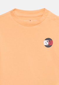 Κοντομάνικο μπλουζάκι με στρογγυλή λαιμόκοψη από βαμβάκι σε ανοιχτό πορτοκαλί χρώμα. Διαθέτει κυκλικό γραφικό λογότυπο στο στήθος με μαύρες, λευκές και κόκκινες αποχρώσεις.