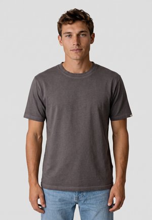 Joven con cabello castaño corto que lleva una camiseta lisa de manga corta de color gris oscuro y vaqueros azul claro, de pie frente a un fondo liso.