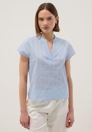 Jeune femme aux cheveux bruns raides portant un chemisier léger texturé bleu clair à manches courtes et un pantalon blanc, debout devant un fond clair uni.