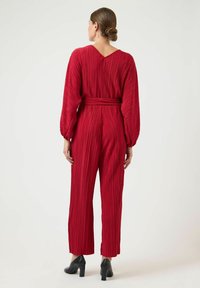 Rød plisseret jumpsuit med lange ærmer og v-hals. Har en taljebånd og vide ben. Afsluttet med sorte hæle.