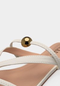 Sandale blanche à fines brides croisées, semelle beige, et perle dorée brillante décorative sur la bride supérieure.