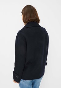 Manteau en laine bleu marine avec un large col et des poignets boutonnés, présentant une coupe droite et une texture lisse, porté sur un denim bleu.