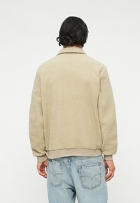 Giacca in pile beige con superficie strutturata, dotata di colletto e polsini a costine, indossata su jeans azzurri chiari. Design minimalista senza marchi visibili.