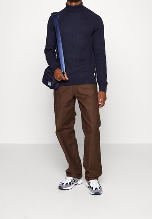 Granatowy sweter z golfem o prążkowanej fakturze, brązowe spodnie i szare oraz białe sneakersy; ciemna torebka na ramię z niebieskim paskiem. Prosty, funkcjonalny design.