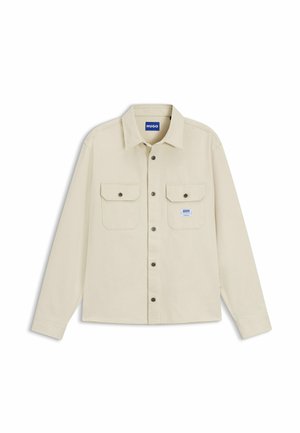 Chemise beige à manches longues avec col, deux poches poitrine avant à rabats boutonnés, et une petite étiquette de marque bleue sur la poche gauche.