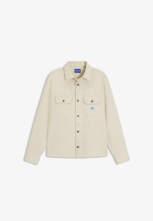 Chemise beige à manches longues avec col, deux poches poitrine avant à rabats boutonnés, et une petite étiquette de marque bleue sur la poche gauche.