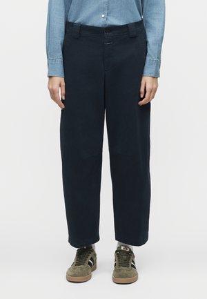 Άτομο που φοράει ναυτικά παντελόνια με φαρδιά μπατζάκια, ανοιχτό μπλε πουκάμισο με κουμπιά, λευκές κάλτσες και ελιάς πράσινα αθλητικά παπούτσια με μαύρες ρίγες.