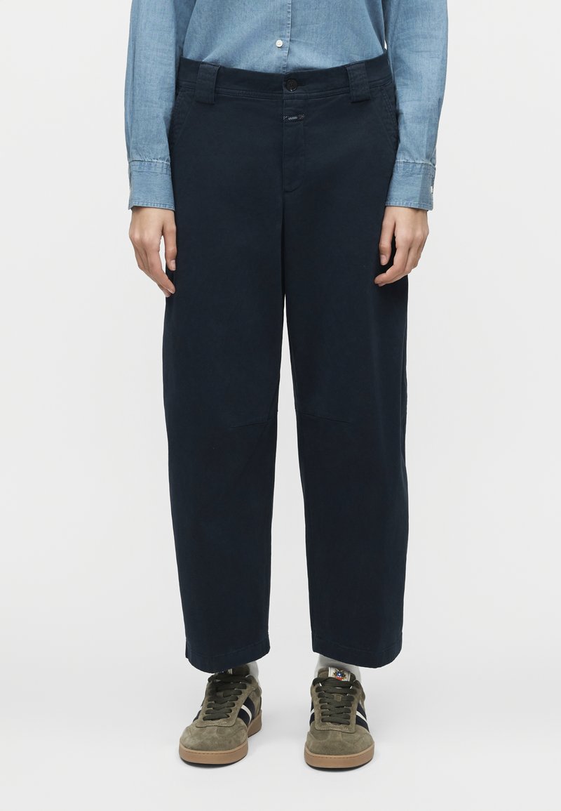 Άτομο που φοράει ναυτικά παντελόνια με φαρδιά μπατζάκια, ανοιχτό μπλε πουκάμισο με κουμπιά, λευκές κάλτσες και ελιάς πράσινα αθλητικά παπούτσια με μαύρες ρίγες.