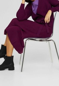 Femme assise sur une chaise transparente, portant une jupe violette, un cardigan assorti ceinturé, un haut violet clair et des bottines noires.