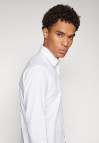Chemise blanche à manches longues avec col boutonné. Texture lisse, design ajusté et léger éclat. Présentée sur un mannequin regardant sur le côté.