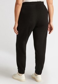 Schwarze Jogginghosen mit einem elastischen Bund und Bündchen, die eine glatte Textur und minimale Details an den Seiten aufweisen. Kombiniert mit weißen Sneakers.