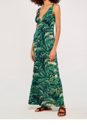 Femme portant une robe longue sans manches à motifs verts et crème avec un décolleté en V profond et des sandales plates marron, debout devant un fond uni.