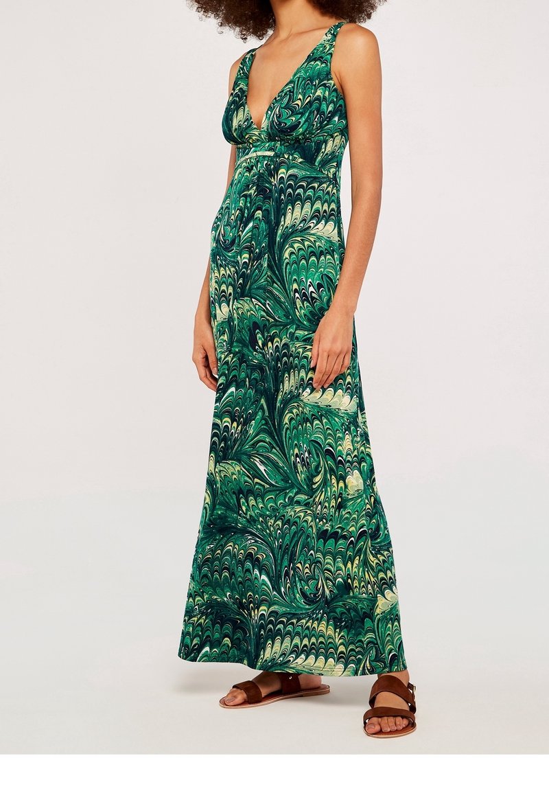 Femme portant une robe longue sans manches à motifs verts et crème avec un décolleté en V profond et des sandales plates marron, debout devant un fond uni.