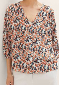 Femme portant un chemisier à col en V avec un imprimé floral orange, bleu et blanc, manches trois-quarts, assorti à un pantalon de couleur claire.