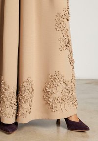 Vestido beige con tela texturizada, adornado con apliques florales en el dobladillo. Combinado con un zapato de tacón alto de color púrpura oscuro.