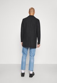 Abrigo de lana negro con corte recto, cuello con muescas y dos bolsillos laterales; llevado sobre pantalones vaqueros de color azul claro y zapatos negros.