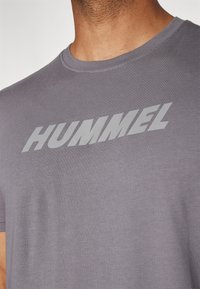 Camiseta de algodón gris con un prominente y audaz logo "HUMMEL" en claro en la parte delantera. Cuello redondo con una textura suave.