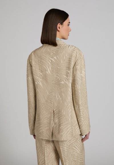Donna con capelli castani lisci indossa un blazer beige e pantaloni coordinati con motivo in rilievo a forma di foglia, in piedi davanti a uno sfondo semplice.
