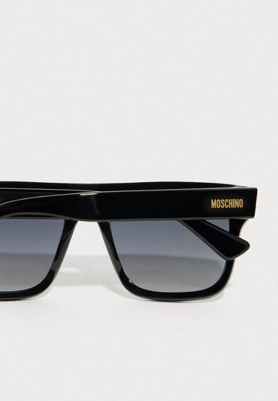 Μαύρα γυαλιά ηλίου με γυαλιστερή φινίρισμα, που διαθέτουν μοναδικό γεωμετρικό σχήμα και ταλαντευόμενους φακούς. Η μάρκα είναι ορατή στο βραχίονα.