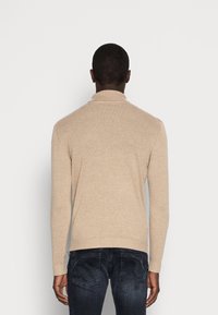 Man som bär en beige långärmad tubtröja och mörka jeans, står vänd mot en vanlig vit bakgrund med avslappnade armar.