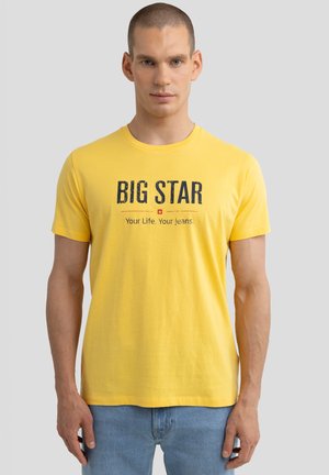 Kollane puuvillane T-särk, millel on julge "BIG STAR" trükk ja allolev loosung, lühikesed varrukad ja ümmargune kaelus. Kandmine siniste teksadega.