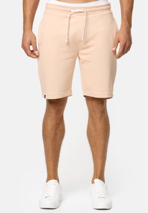 Mann trägt hellbeige Shorts mit Kordelzug und weiße Turnschuhe, steht vor einfachem Hintergrund.
