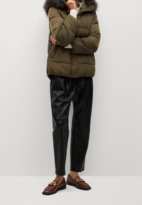 Osoba ubrana w oliwkową kurtkę puchową z futrzanym kapturem, czarne skórzane spodnie oraz brązowe mokasyny z detalem w postaci złotego łańcucha, stojąca przy białej ścianie.