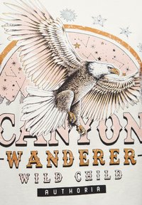 Kreemist t-särki, millel on suur lennukäed, peene detailiga ja graafiline disain, millel on sõnad "CANYON WANDERER" segafontides.