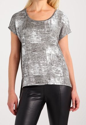 Woman wearing a short-sleeve, silver metallic textured top with a rounded neckline and black leather pants against a plain background.