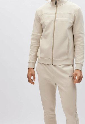 Homme portant une veste beige zippée et un pantalon assorti avec le logo BOSS sur la poitrine et la cuisse, debout devant un fond uni.