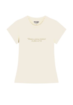 Femme portant une veste crème avec un bord noir sur un t-shirt clair et un pantalon noir, regardant vers le bas avec les mains près des hanches.