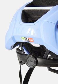 Kask kask rowerowy w jasnoniebieskim kolorze. Posiada regulowane paski, otwory wentylacyjne oraz czarny system mocujący; zawiera etykiety certyfikacji bezpieczeństwa.