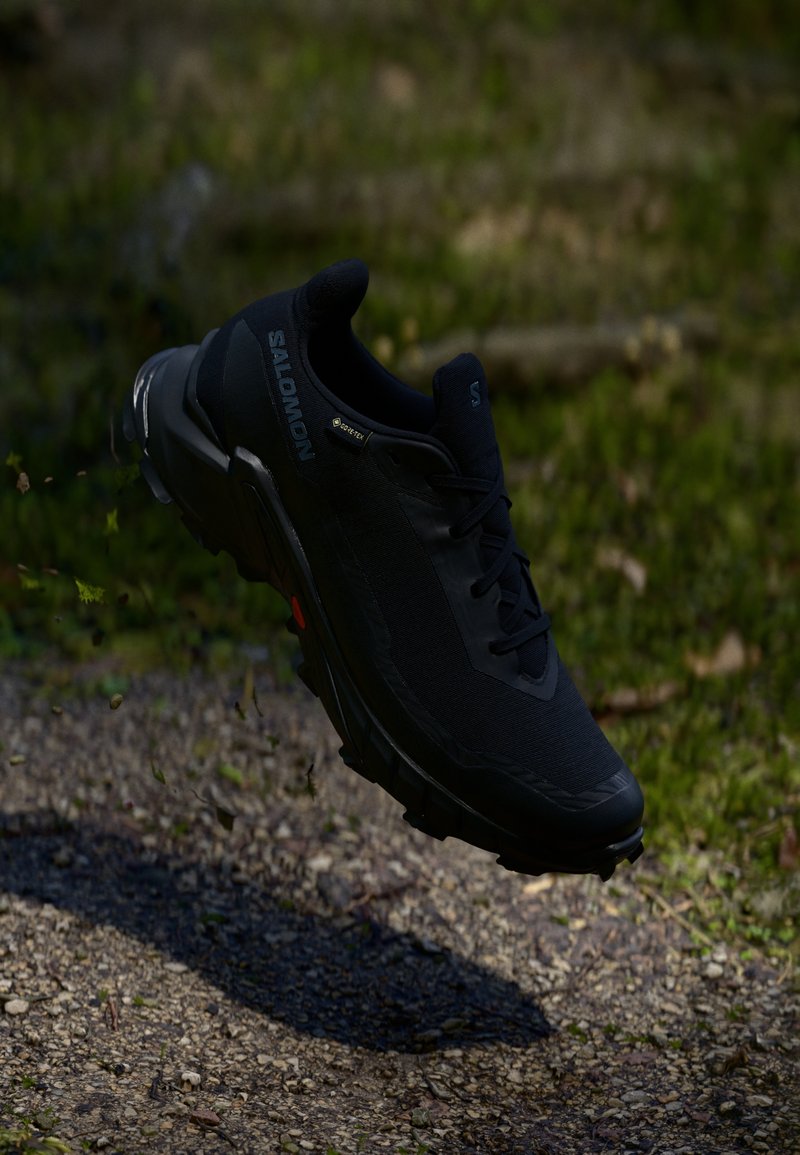 Chaussure de trail running noire avec un dessus respirant, un tissu texturé et une semelle en caoutchouc. Elle présente un profil bas, un système de laçage et un logo discret.