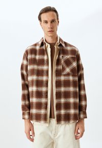 Camicia flanella a quadri marrone e crema con chiusura a bottoni, tasca sul petto e vestibilità comoda, indossata sopra un top beige.