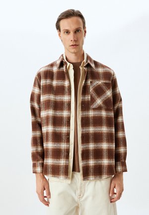 Camisa de franela de cuadros marrón y crema con cierre de botones, bolsillo en el pecho y corte relajado, superpuesta sobre una camiseta beige.