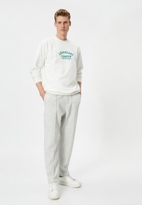 Sudadera blanca de cuello redondo con texto verde "LEYENDA CAMPEONA", combinada con pantalones gris claro con pliegues y zapatillas blancas.
