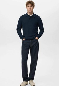 Granatowa koszulka polo z długim rękawem i kołnierzykiem, zestawiona z ciemnoniebieskimi dżinsami i beżowymi butami, stojąca na neutralnym tle.