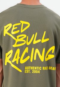 Alyvučių žalumo medvilninis marškinėliai su išraiškingu geltonu tekstu: "RED BULL RACING" ir "AUTHENTIC RACE GEAR EST. 2004" skirtingomis šrifto stiliais.