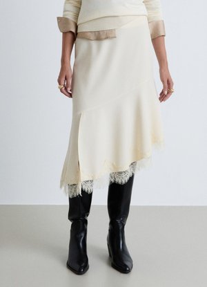 Donna con gonna asimmetrica color crema con bordatura in pizzo sopra stivali neri con tacco e top beige a strati con maniche arrotolate.