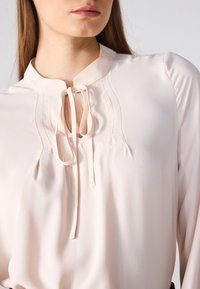 Blusa de color rosa claro con cuello mao, detalles pleat y un lazo en el escote. La tela parece suave y ligeramente brillante.