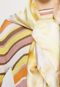 Écharpe en soie dans des nuances de jaune doux, présentant une texture lisse. Portée drapée sur un haut en maille rayé multicolore avec des rayures orange, rose et marron.