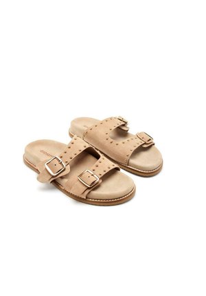 Chanclas de ante en beige claro con dos correas anchas, hebillas en tono dorado y acentos de tachuelas. Suela plana con un acabado texturizado.
