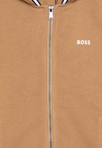 Beżowa bluza z kapturem na zamek błyskawiczny, z srebrnym zamkiem i czarno-białą paskowaną stójką, z logo "BOSS" na lewej piersi.