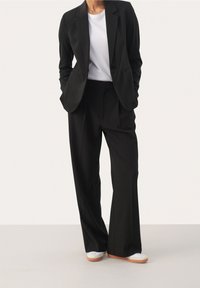 Veste de costume noire à revers crantés, associée à un pantalon large noir et un t-shirt blanc, complétés par des baskets blanches. Texture de tissu lisse.