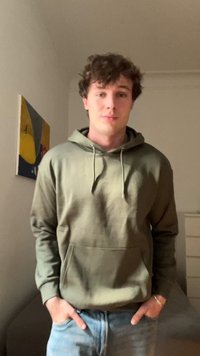 Jonge man met krullend haar, gekleed in een olijfgroene hoodie en jeans, staande binnen met zijn handen in de zakken, abstract schilderij op de muur achter hem.