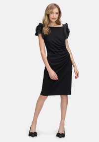 Schwarzes Kleid mit gerafften Details, kurzen Puffärmeln und geradem Schnitt. Getragen mit schwarzen Schuhen mit Schleifenakzenten. Glatte Stofftextur.