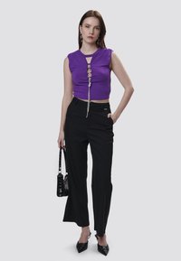 Top corto senza maniche di colore viola con lacci frontali, abbinato a pantaloni neri a vita alta; modella che tiene una piccola borsa nera e indossa scarpe a punta.