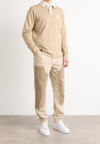 Beige långärmad polo med kontrasterande krage, matchad med beige byxor som har kordflorsepatchar; modellen bär vita sneakers.