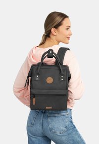 Sac à dos noir de forme rectangulaire, en matériau synthétique, avec une poignée supérieure, des bretelles ajustables, des poches zippées et un accent logo.