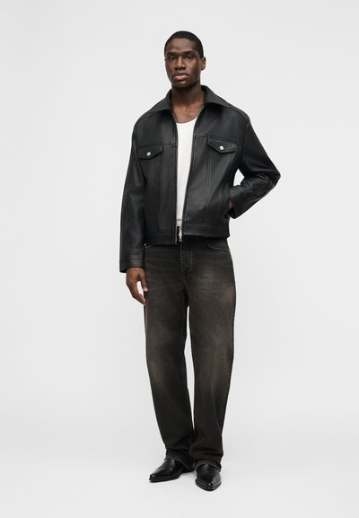 Άνδρας που στέκεται φορώντας μαύρο δερμάτινο μπουφάν, λευκό πουκάμισο, ξεβαμμένα μαύρα τζιν και μαύρα παπούτσια με μυτερή μύτη μπροστά σε απλό φόντο.